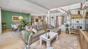A spacious living room with a grey sofa, a coffee table, and a dining area with chairs and a table. There is a staircase leading to an upper floor. - Luxury Villa in Serene Location w/ Pool & Private Garden (Quarteira)