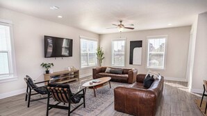 A living room with a brown leather sofa, a coffee table, and a TV mounted on the wall. There are windows with blinds, a ceiling fan, and a bookshelf. - Beach Retreat Complex w/Pool - Sleeps 25! (Jacksonville Beach)