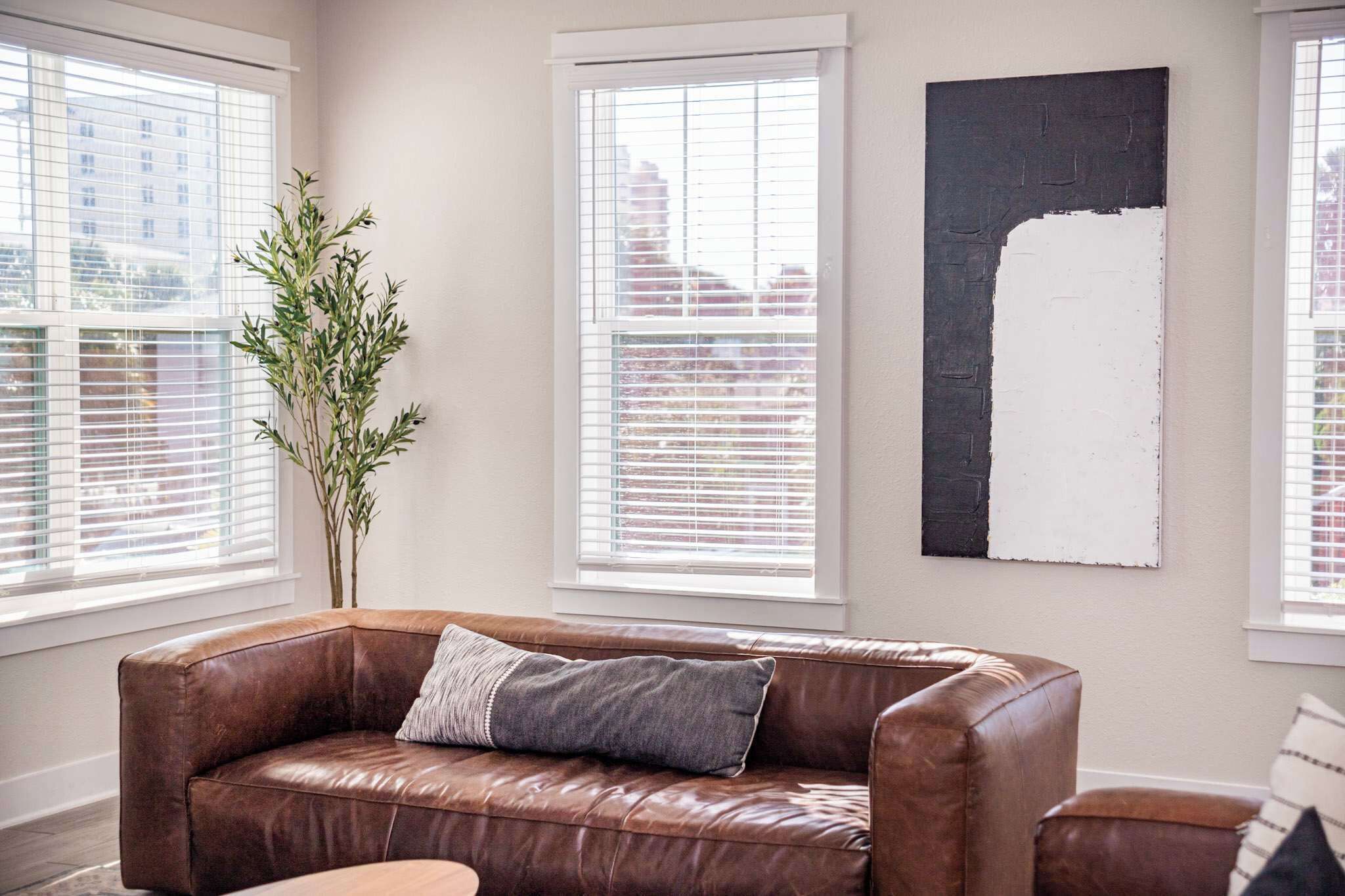 Een kamer met een bruine leren bank, een groot raam met zonwering, een hoge plant en een groot abstract schilderij aan de muur.