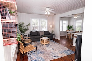 A spacious living room with a dark grey sofa, a round coffee table, and a blue patterned rug. There are two windows with white blinds, a ceiling fan, and a dining area visible in the background.