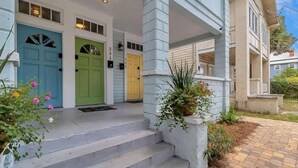 Une maison à deux étages avec un porche à l’avant, trois portes (bleue, verte et jaune) et un chemin en briques menant à l’entrée.