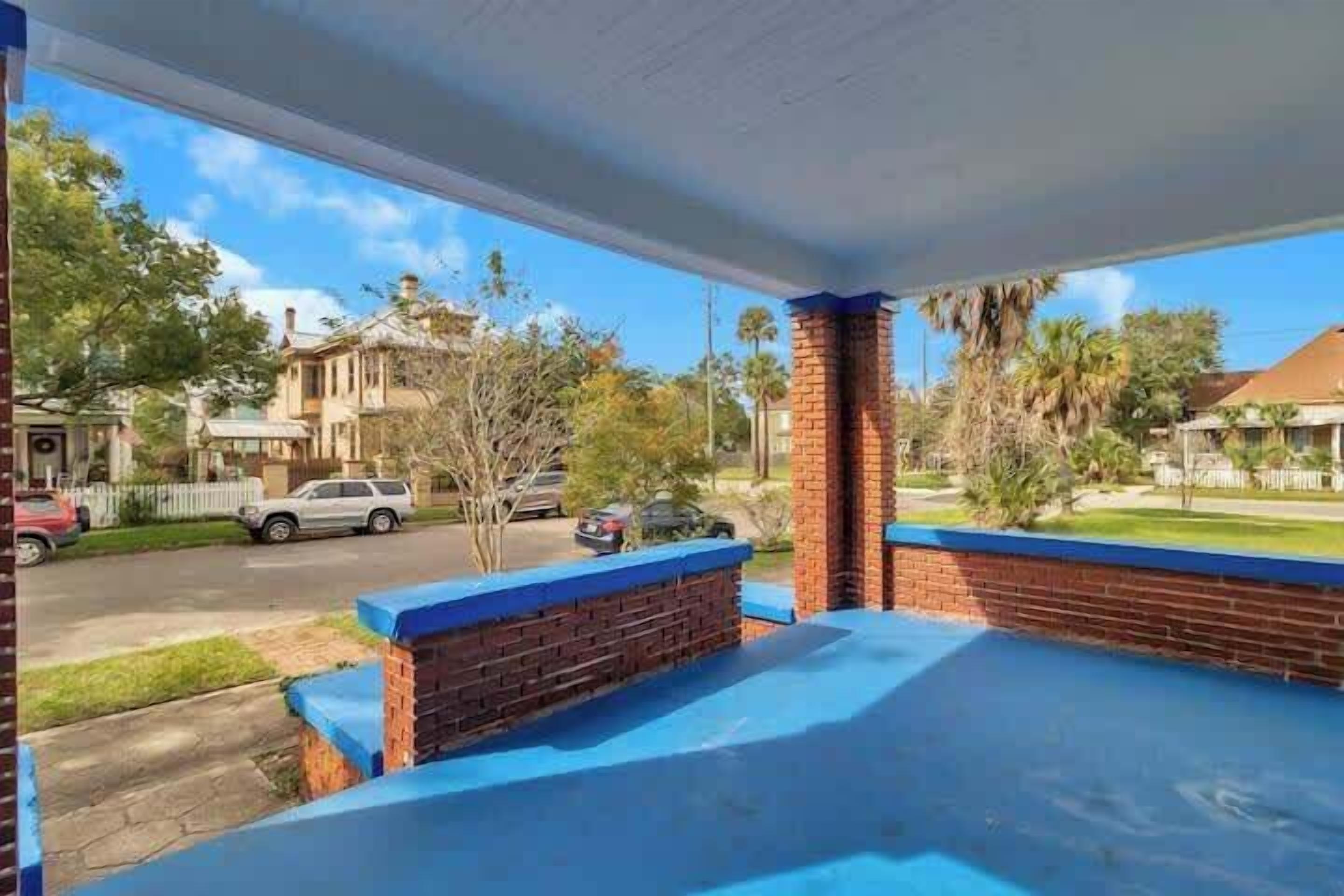 Un espacio al aire libre cubierto, con pared de ladrillo y detalles azules, que da vista a una calle con autos estacionados y casas.