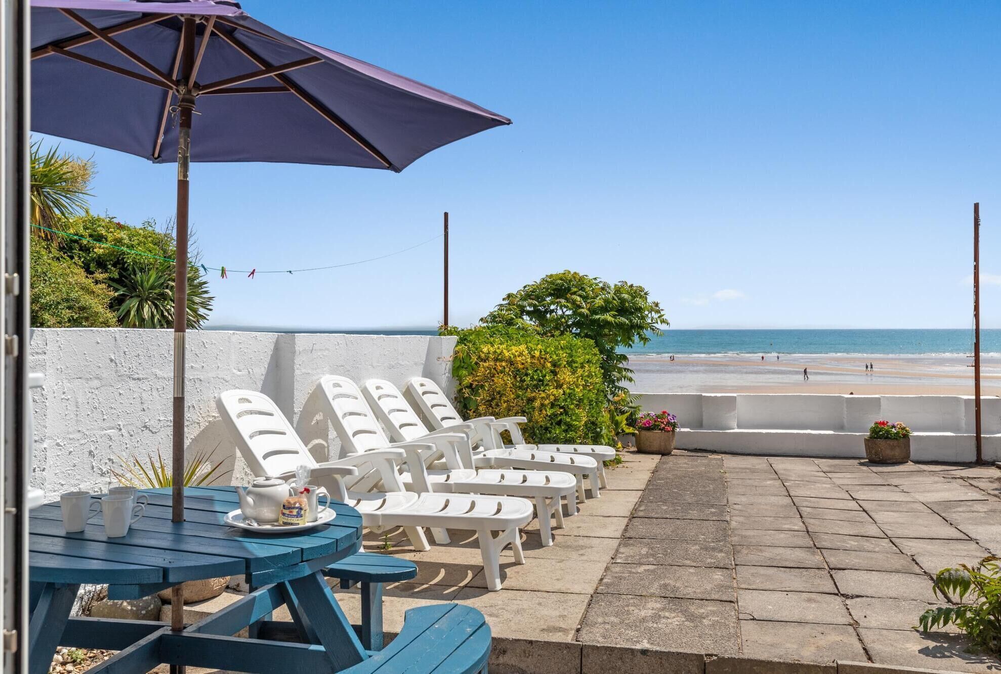 A patio with white lounge chairs and a table with a teapot and cups, overlooking a beach.