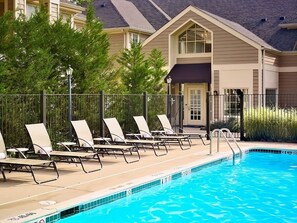 Une piscine dotée de chaises longues et entourée d’une clôture, le tout bordé de verdure et adossé à un bâtiment au toit en pente.