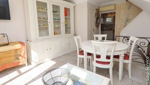 A dining area with a white table and chairs, a white cabinet with glass doors, and a kitchen visible through an open door.