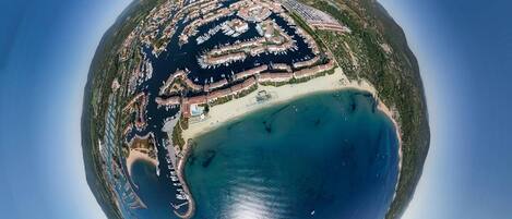 Vista aérea de un complejo turístico costero con puerto deportivo, playa de arena y abundante vegetación.