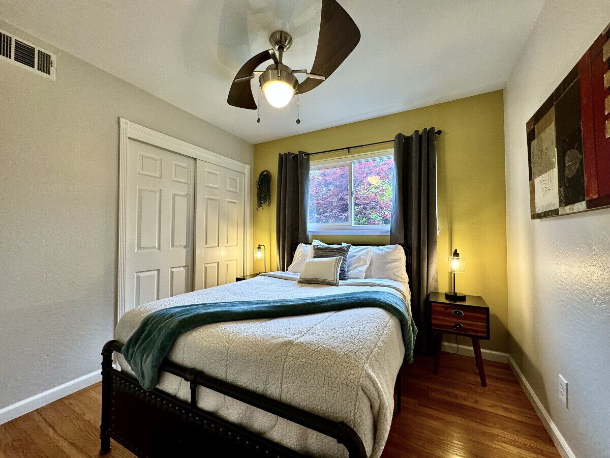 A bedroom with a bed, a ceiling fan, a window with curtains, a nightstand, and a wall-mounted lamp.
