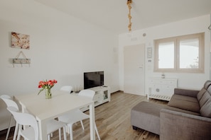 A modern, well-lit room with a dining table, white chairs, a grey sofa, a TV, and a wooden floor.