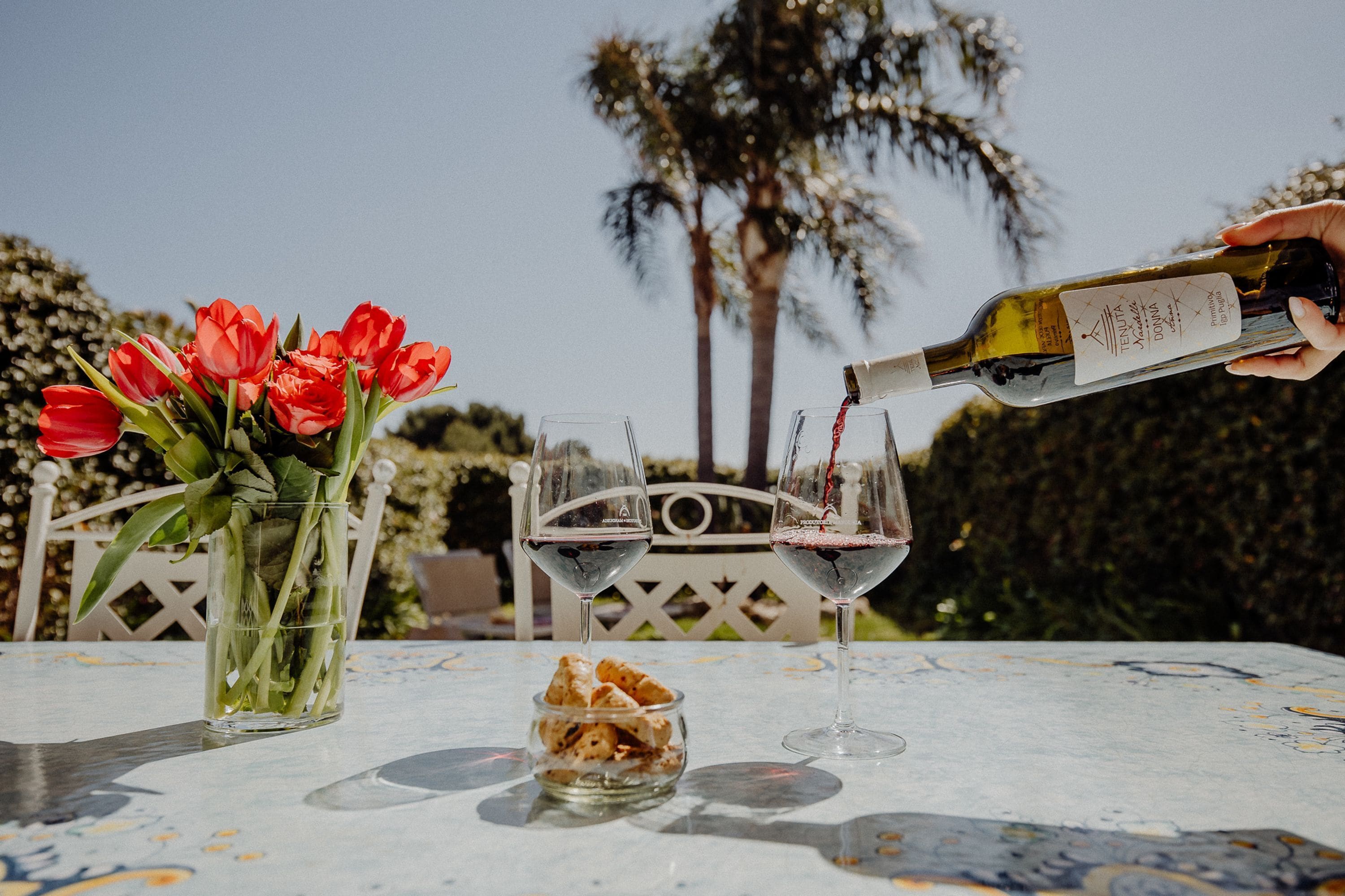 A table with two wine glasses, a bottle of wine, a vase with red flowers, and a bowl of snacks.