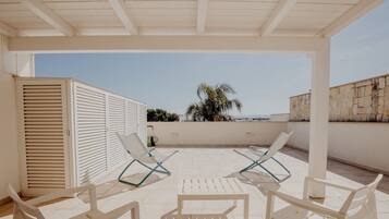 A covered outdoor seating area with two lounge chairs and a table, overlooking a palm tree and a stone wall.