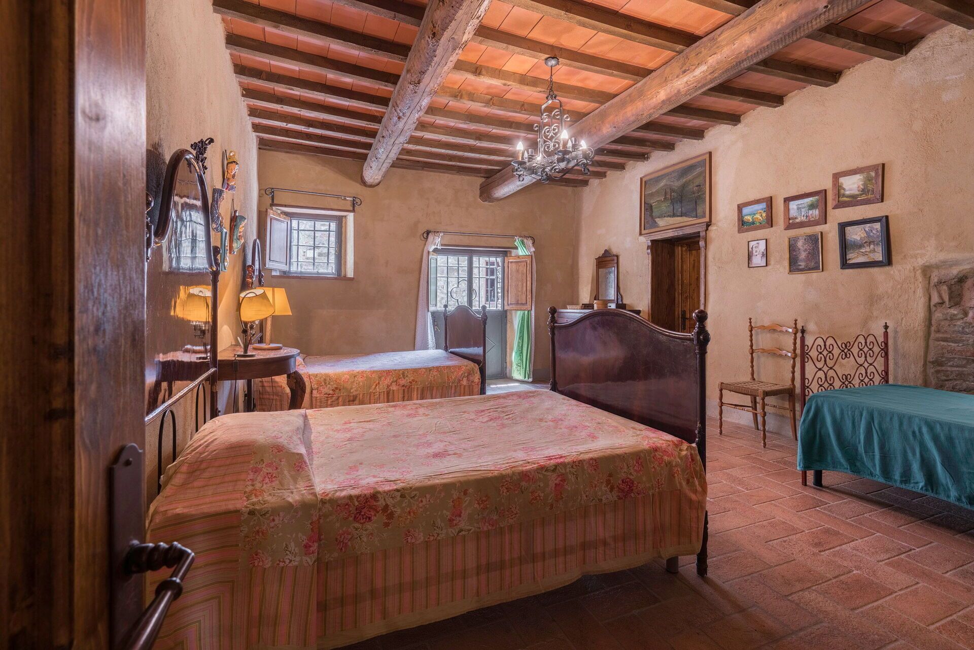 A rustic room with a wooden ceiling, a bed with floral bedding, a wooden chair, and a table with a lamp.