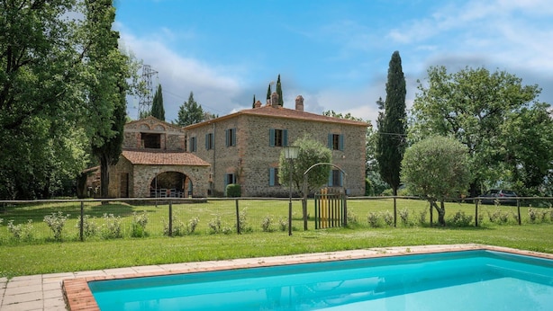 Una villa in pietra con piscina, immersa in un giardino recintato e alberi alti.
