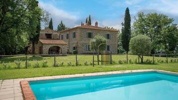 A stone villa with a swimming pool, surrounded by a fenced garden and tall trees.
