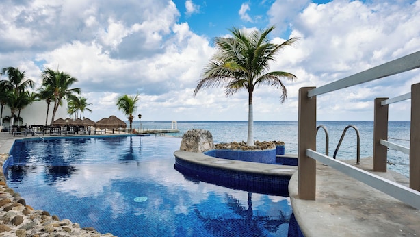 A pool area with a view of the ocean, palm trees, and a thatched-roof structure. - 
7th-Floor Oceanfront Oasis: 4BR/4BA, Dive Pier & Walk to Town! (San Miguel de Cozumel)