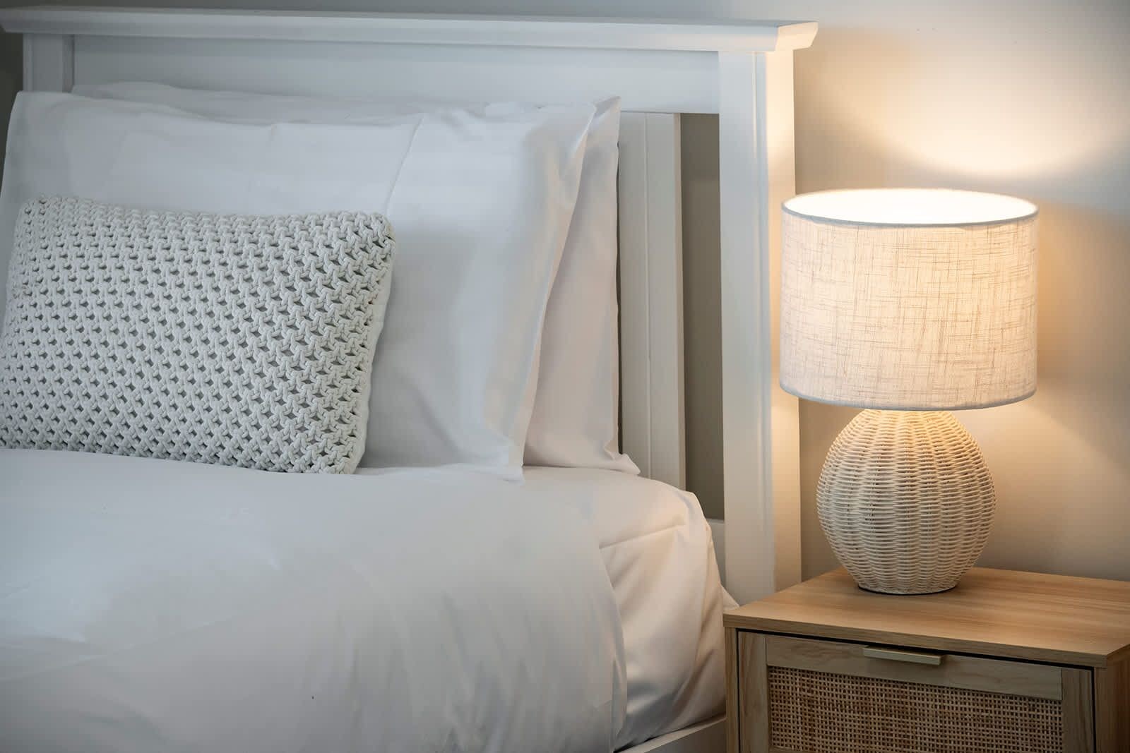 A bed with white linens and a textured pillow, a bedside lamp with a woven base, and a wooden nightstand with a drawer.