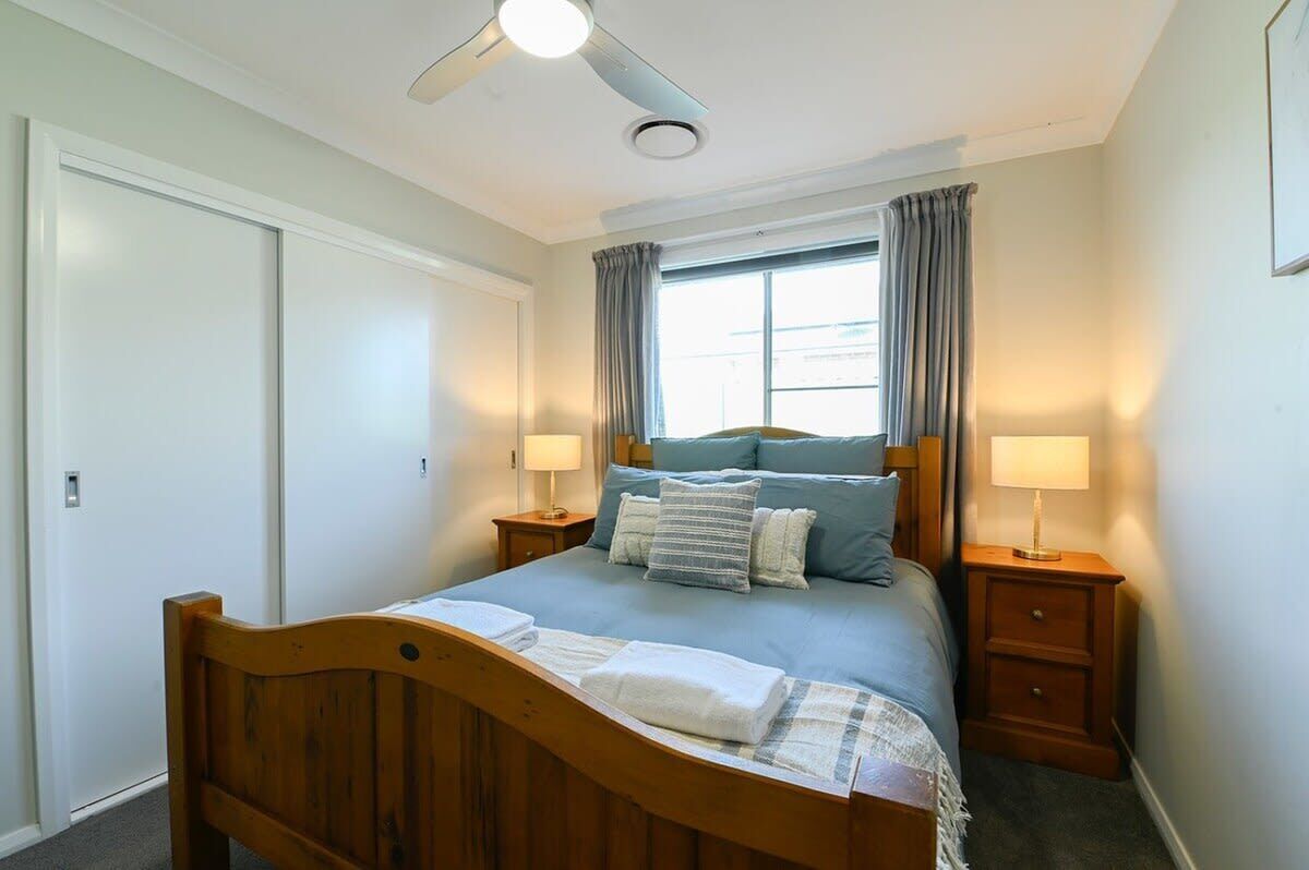 Une chambre à coucher avec un lit en bois, deux tables de chevet avec des lampes, un ventilateur de plafond, une fenêtre avec des rideaux et une porte.