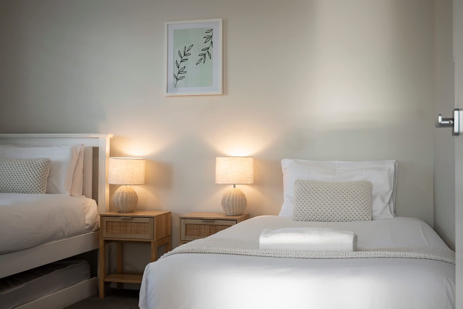 Ein Hotelzimmer mit zwei Betten, jeweils mit Nachttisch und Lampe. Darüber hängt ein gerahmtes Kunstwerk an der Wand.