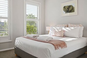 A bedroom with a bed, pillows, a blanket, a framed picture on the wall, and windows with blinds.