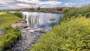 A river flowing through a landscaped area with grass and reeds, leading to a row of modern buildings with large windows and a flat roof. - 9th Hole Clubhouse Stroll (St. George)