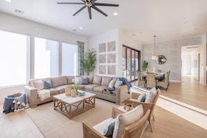Uma sala de estar espaçosa com sofás, mesa de centro e cadeiras de vime. Há uma área de jantar com mesa e cadeiras, uma janela grande e um ventilador de teto.