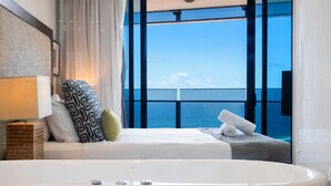 Une salle de bain moderne avec une grande baignoire, une fenêtre donnant sur la mer et un lit bien fait, agrémenté d’oreillers.