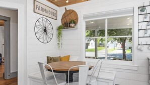 Een eetruimte met een houten tafel en witte stoelen, een groot raam met uitzicht naar buiten, en een wandplank met planten.