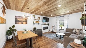 Een moderne woonkamer met een houten plafond, een eethoek met een houten tafel en metalen stoelen, een grijze bank en een aan de muur bevestigde televisie.