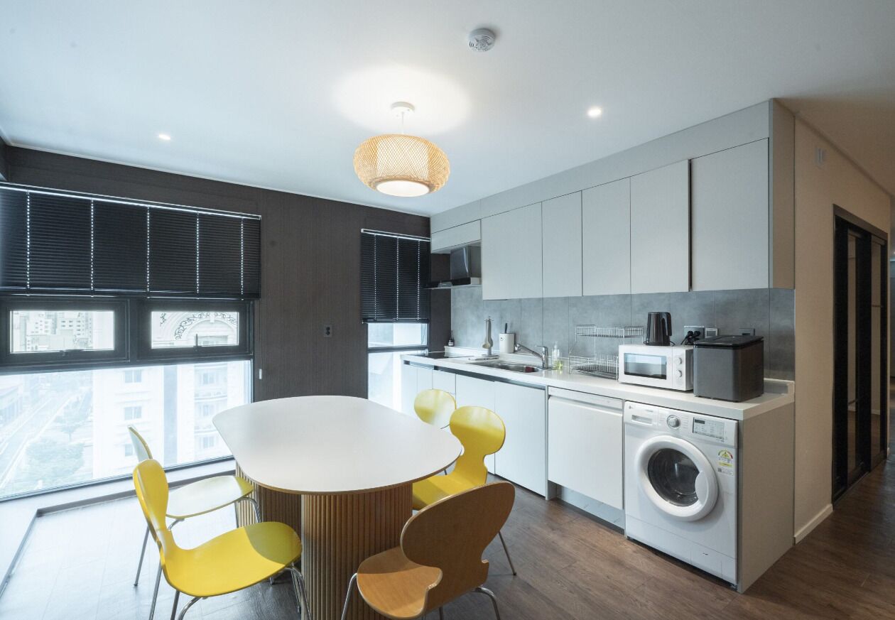 A modern kitchen with a round dining table, yellow chairs, a washing machine, and a large window.