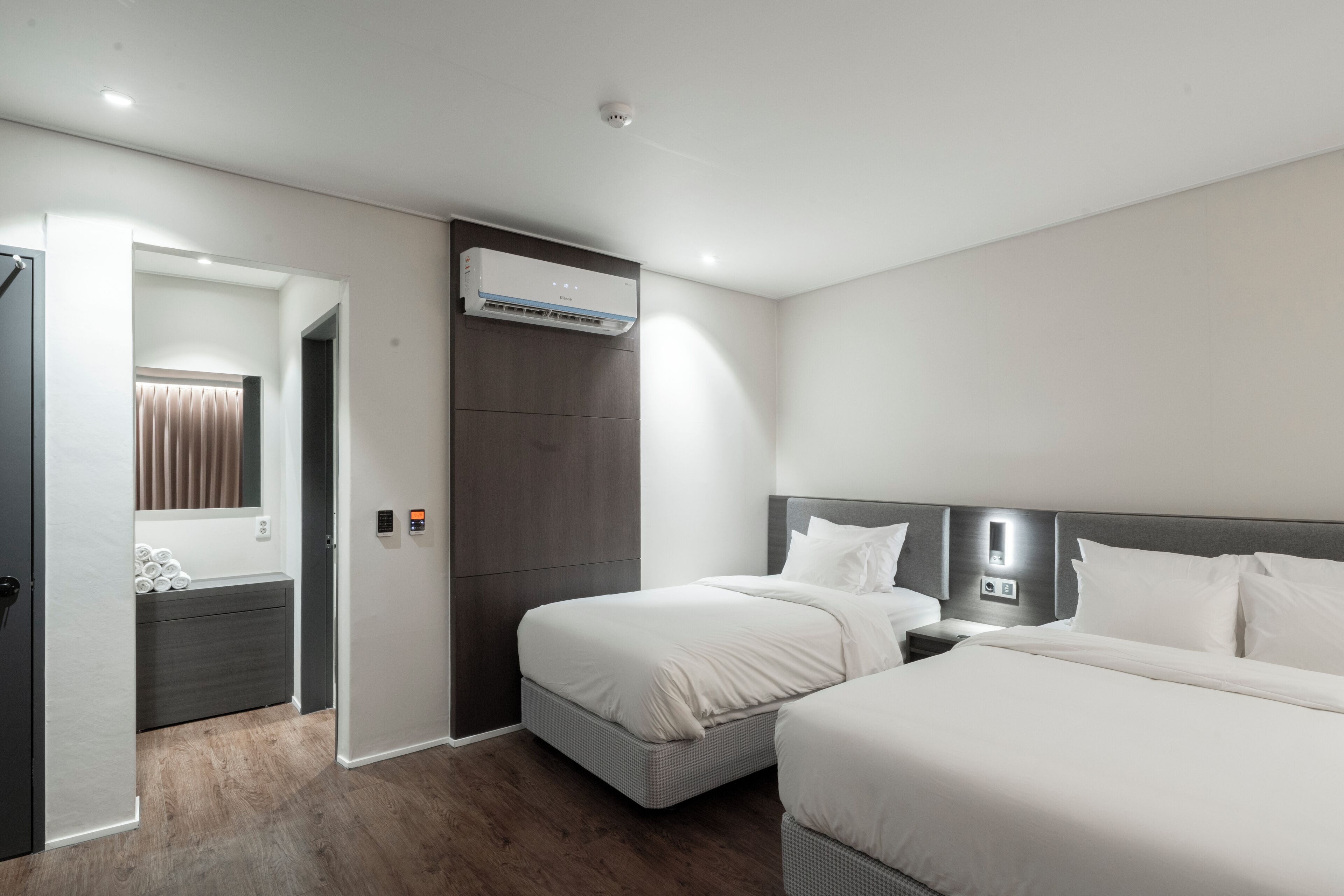 Una habitación de hotel con dos camas, un armario, un espejo, un equipo de aire acondicionado y una mesita de noche con lámpara.