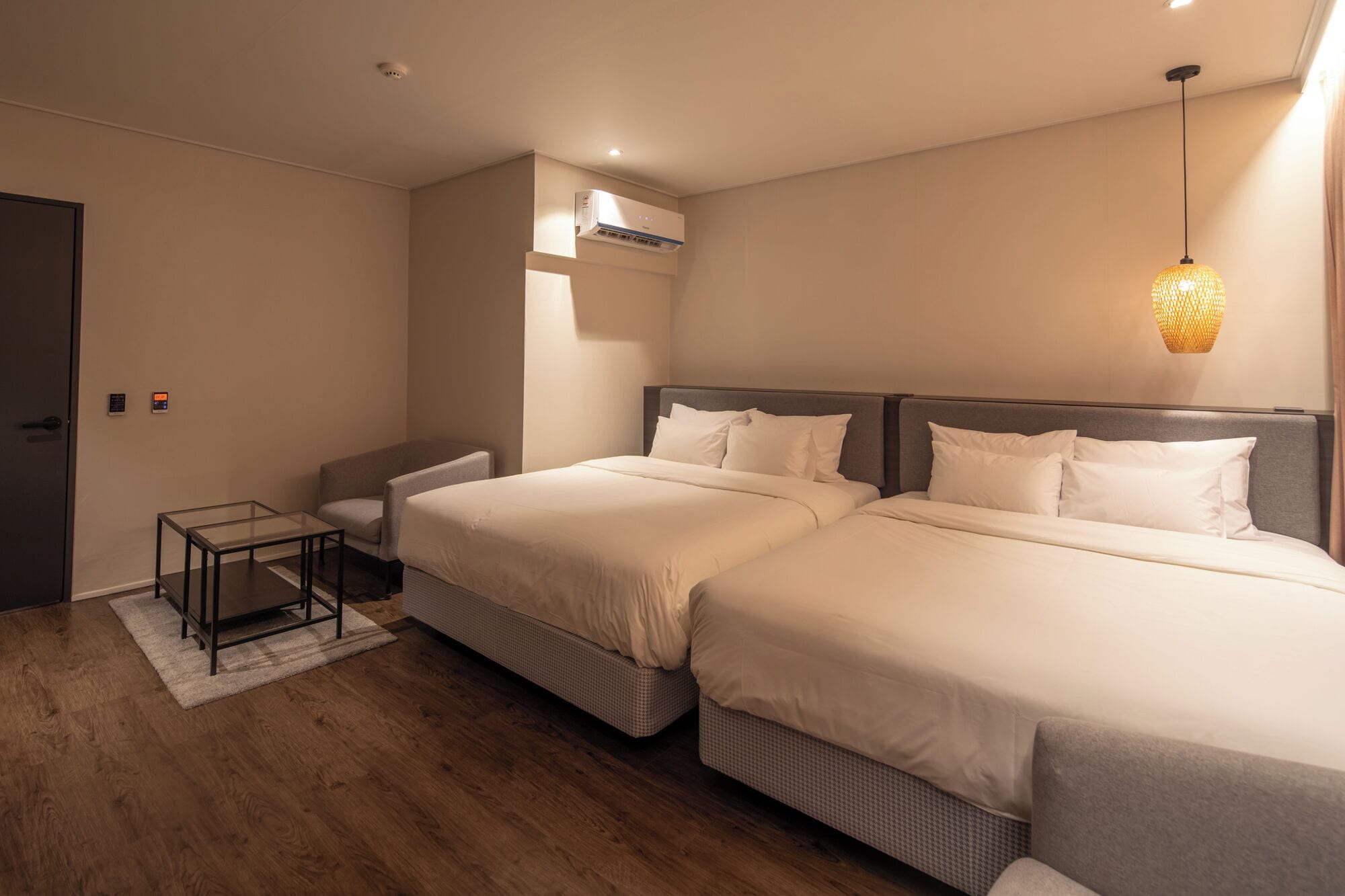 A hotel room with two beds, a small table, a chair, a standing heater, and a wall-mounted air conditioner.