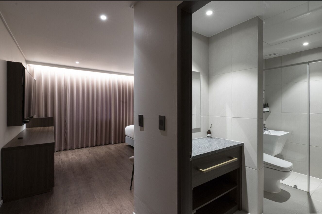 A modern hotel room with a wooden desk, a bed, a bathroom with a glass shower, and a large mirror.