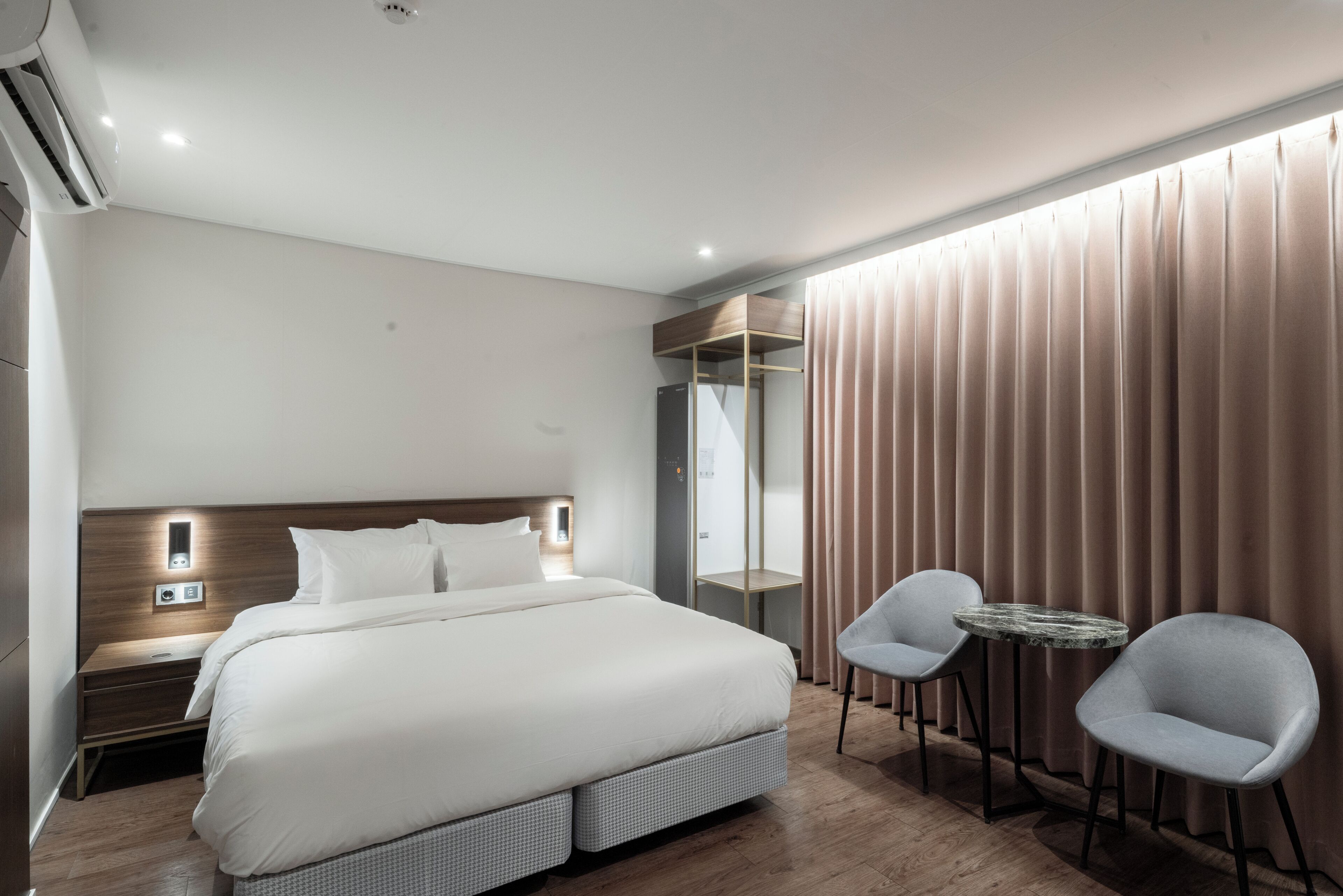 A modern hotel room with a large bed, bedside tables, a wardrobe, a mirror, and two chairs.