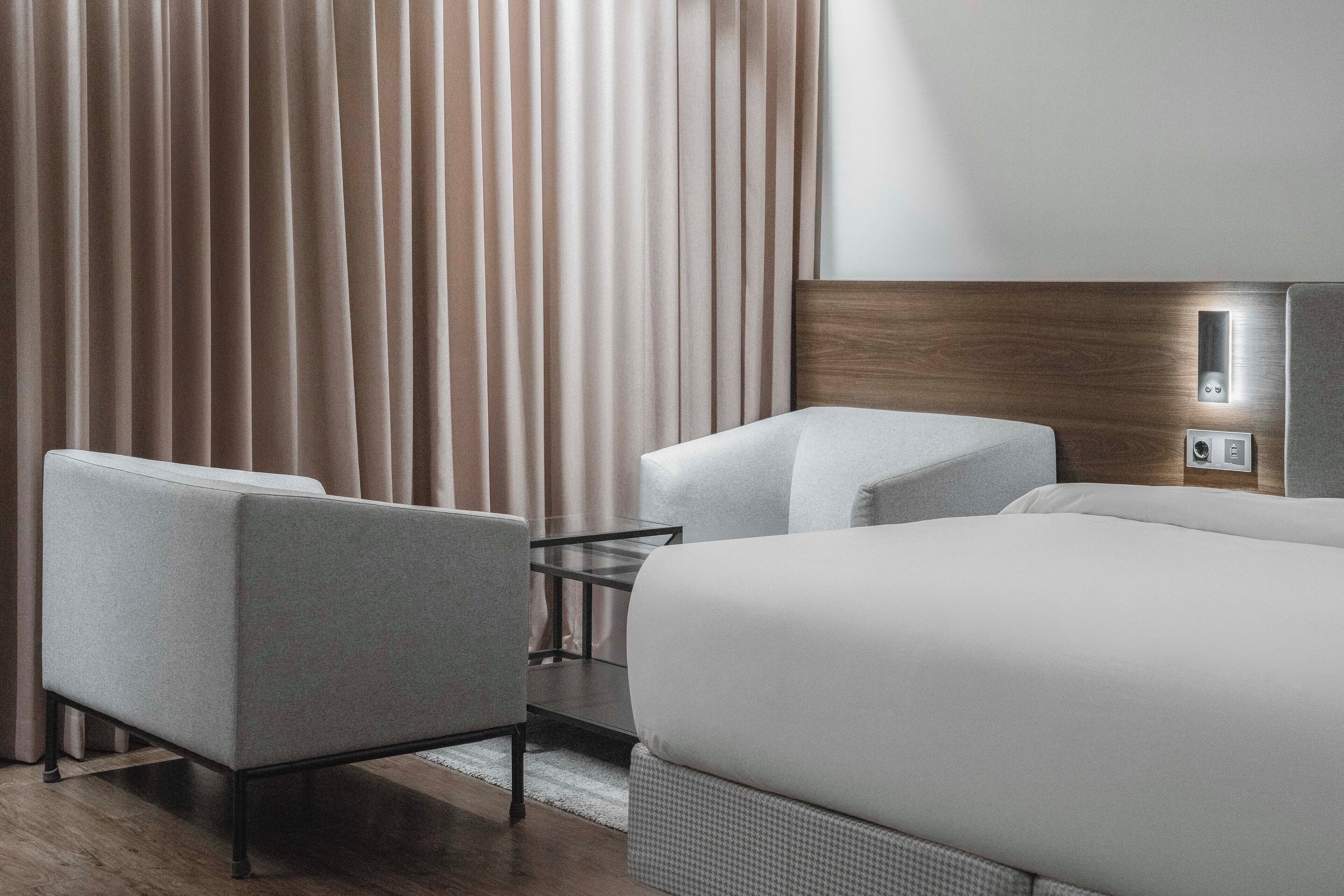 A modern hotel room with a large bed, two armchairs, a small table, and a wooden headboard.