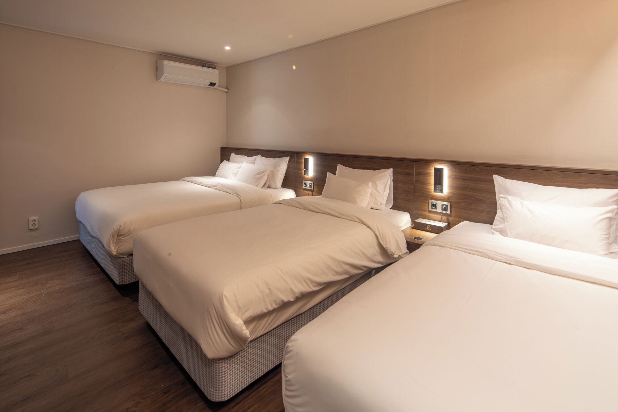 Habitación de hotel con dos camas, una mesita de noche con lámpara y un equipo de aire acondicionado en la pared.