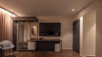 Habitación de hotel moderna con piso de madera, escritorio con televisor de pantalla plana, armario con espejo y zona de estar con una silla.