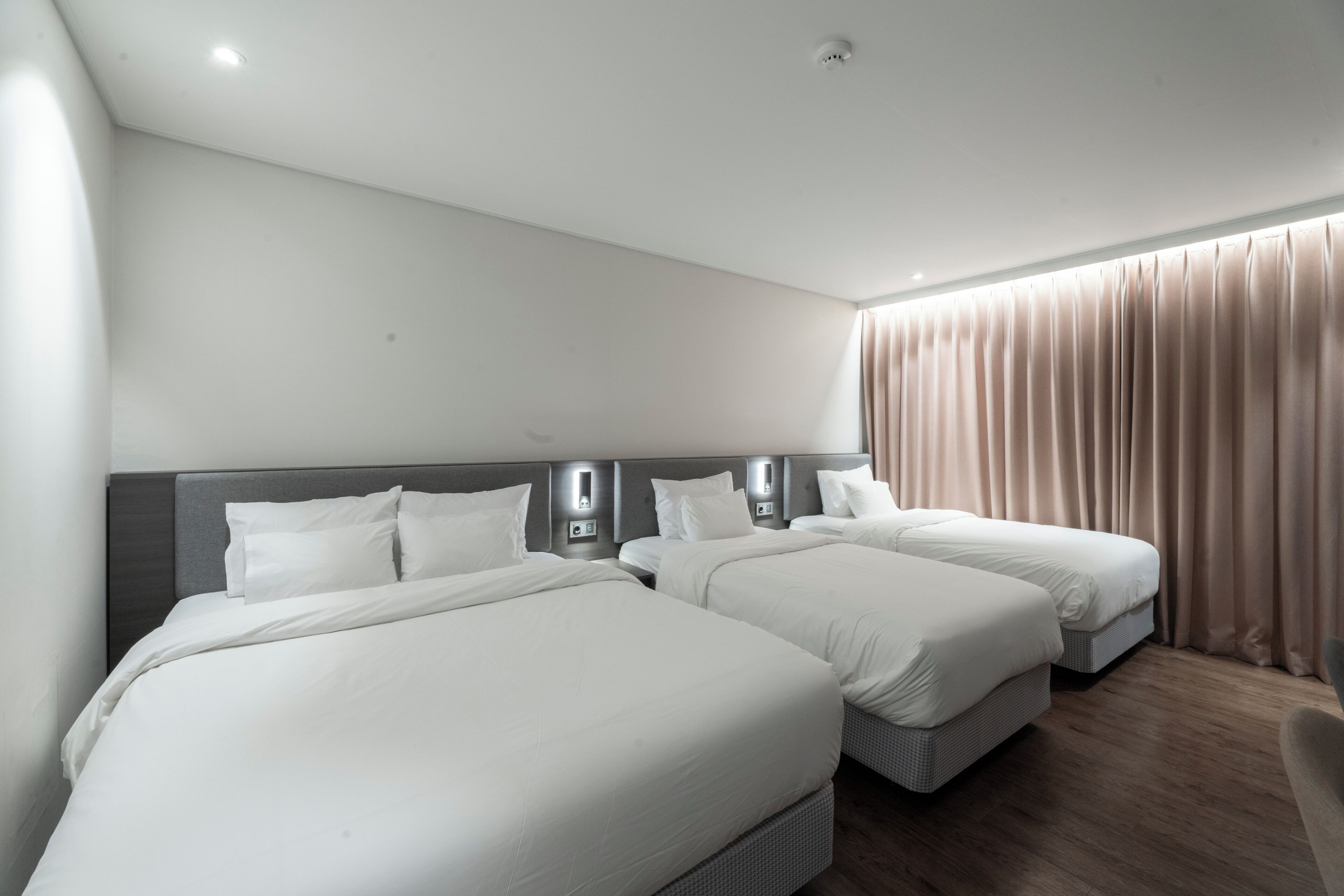 A hotel room with two beds, a headboard with integrated lighting, a large window with curtains, and a wooden floor.