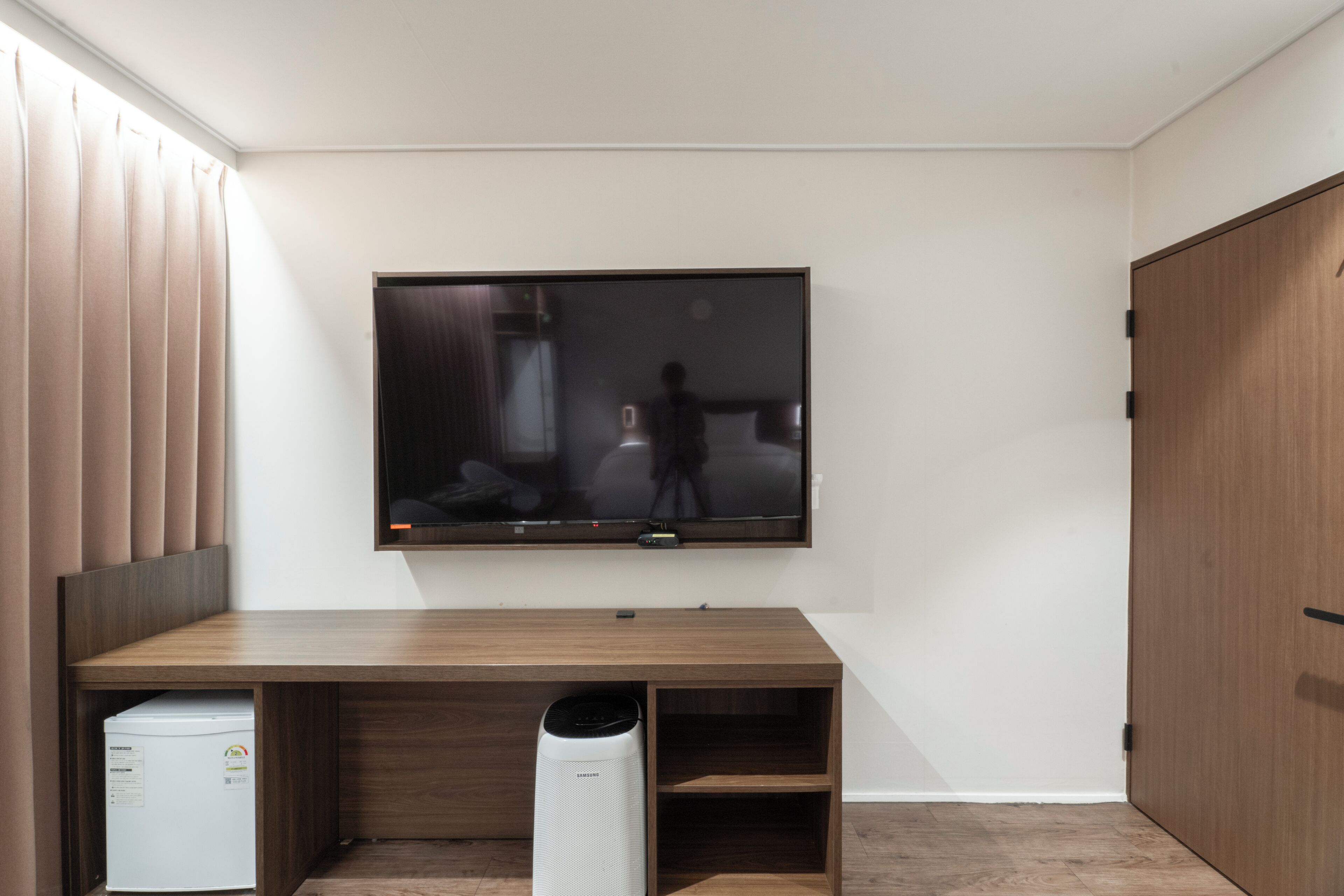 A room with a flat-screen TV mounted on the wall, a wooden desk, a white appliance, and a tall white device on the floor.