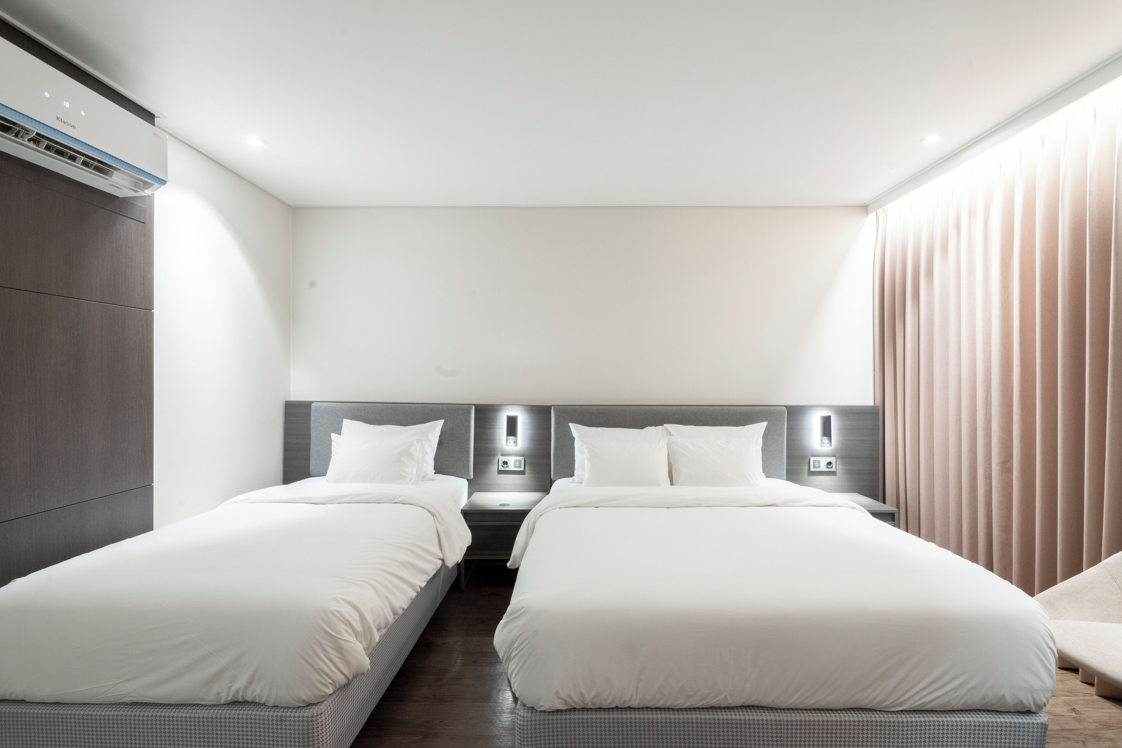 A modern hotel room with two single beds, a built-in air conditioning unit, bedside tables, and a large window with curtains.