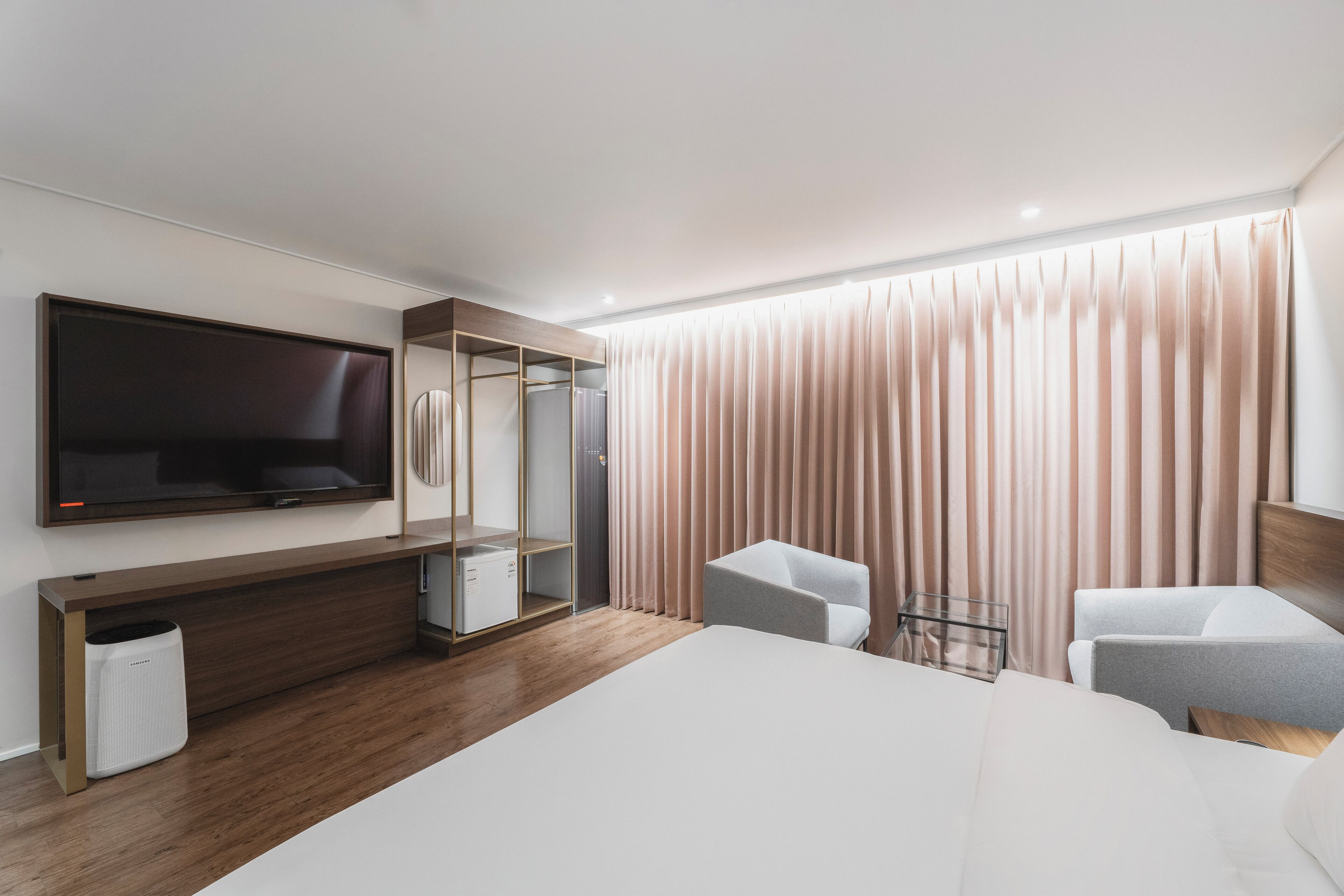 A modern hotel room with a large bed, a TV mounted on the wall, a seating area with two chairs, and a wardrobe with a mirror.