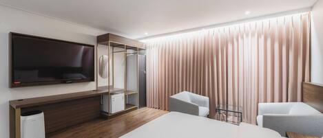 A modern hotel room with a large bed, a TV mounted on the wall, a seating area with two chairs, and a wardrobe with a mirror.