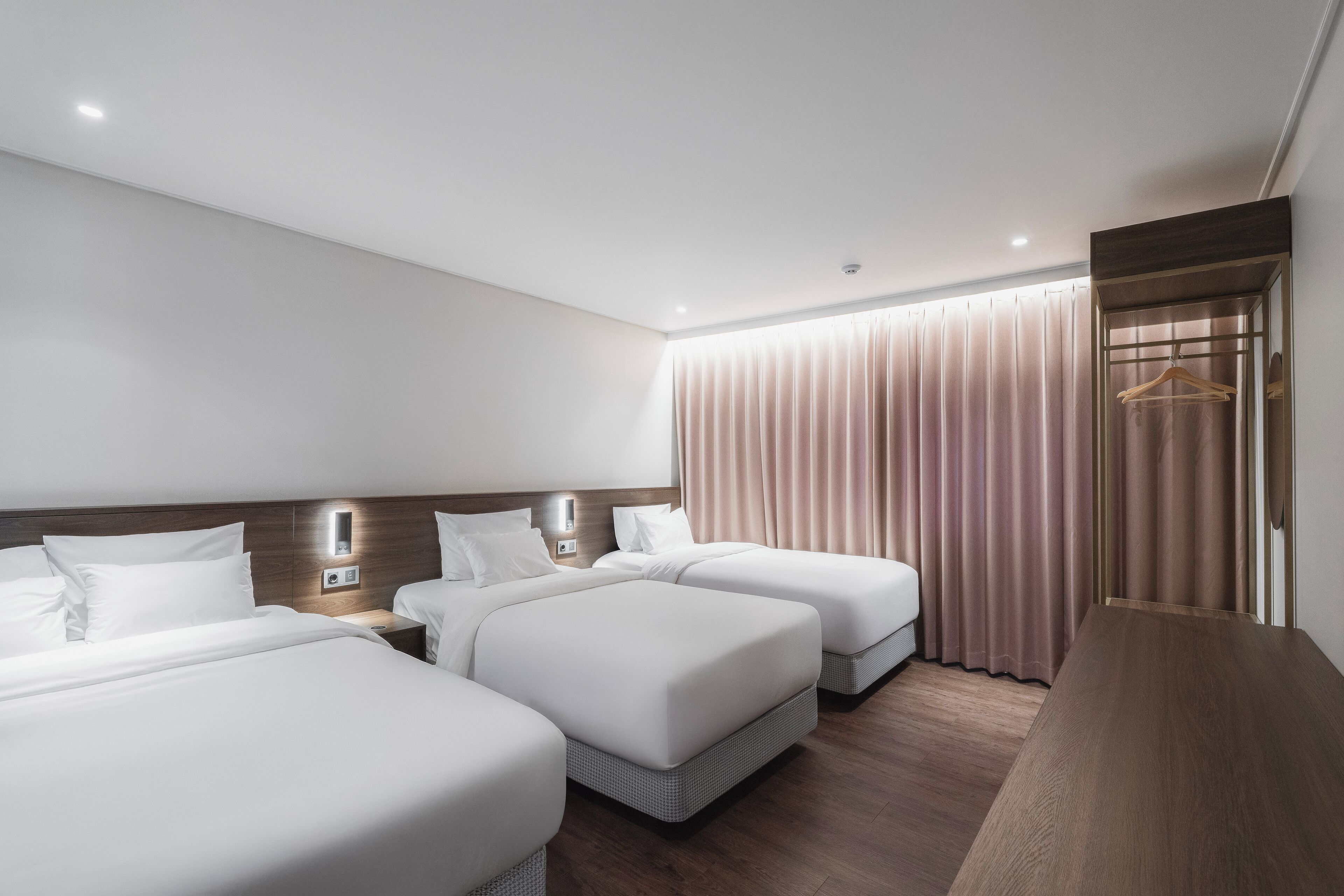 Habitación de hotel con dos camas, cabecero de madera, mesita de noche, perchero y un amplio ventanal con cortinas.