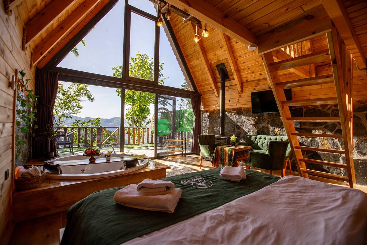 A cozy wooden cabin with a large window, a wooden bed, a small table, and a chair.