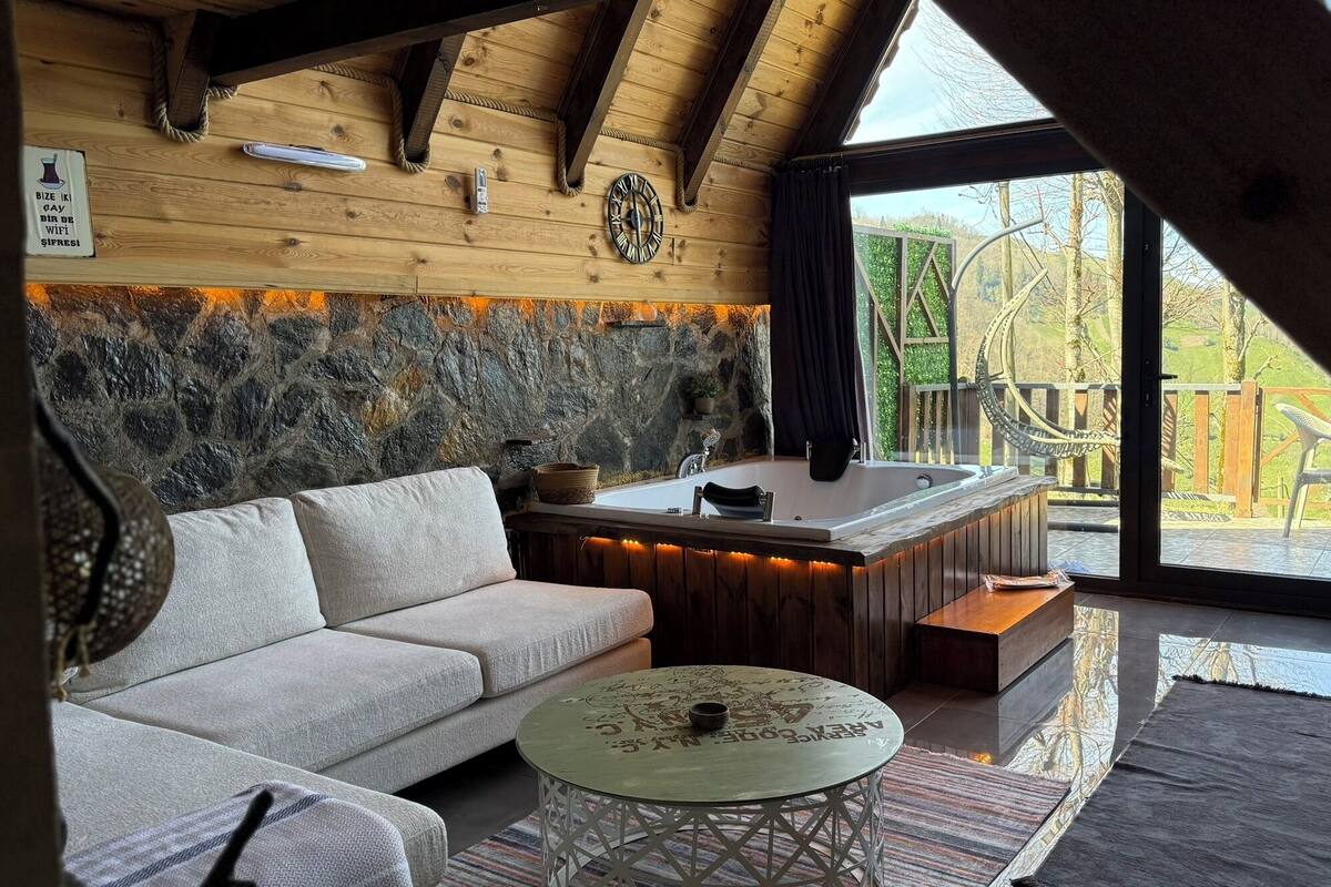 A cozy cabin interior with a stone wall, wooden ceiling, a sofa, a round table, and a view of the outdoors.