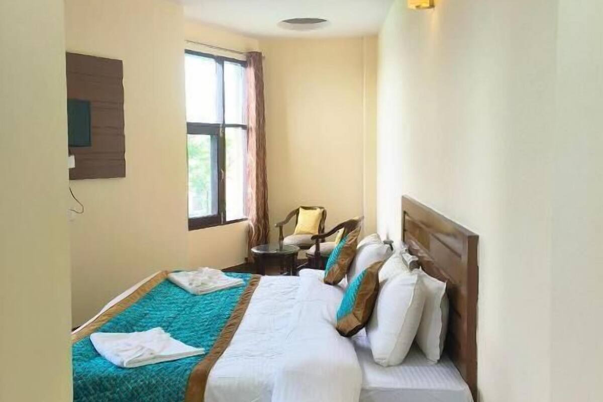 A hotel room with a bed, a small table, a chair, a window, and a wall-mounted light fixture.