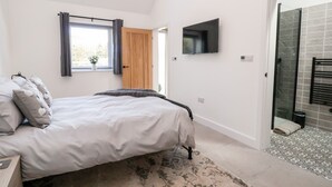 Un dormitorio con cama, ventana con cortinas, televisor en la pared y zona de ducha con piso de baldosas.