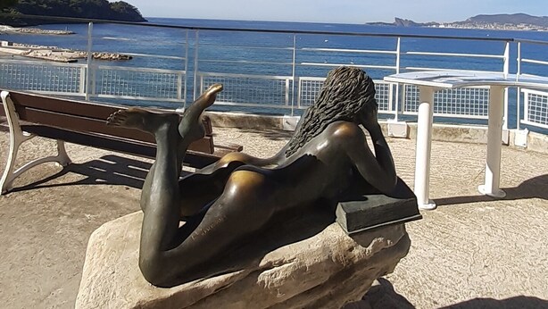 En skulptur som föreställer en liggande figur på en stenplattform, med utsikt över havet och avlägsna kullar.