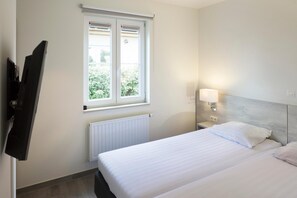 A hotel room with a bed, a nightstand with a lamp, a window with blinds, and a TV mounted on the wall.