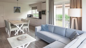 Ein modernes Wohnzimmer mit grauem Sofa, einem Esstisch mit Stühlen, einer Küchenzeile mit Mikrowelle und einem großen Fenster mit Vorhängen.