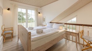 A bedroom with a wooden bed, a desk, and a chair. There are windows with curtains and a view of greenery outside. - Bungalow by the Sea with Sauna in Germany (Sierksdorf)