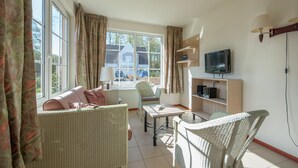A cozy living room with wicker furniture, a television on a wooden shelf, and large windows with curtains.